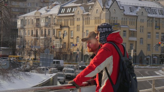 Two Men Are Talking Steam Comes Out of Mouth Standing on a Bridge Winter Vintage Building is in front oa People Man in Red Jacket in Fur Hat Man in Cap