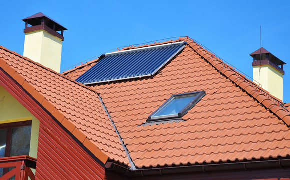 Energy efficiency concept. Closeup of solar water panel heating on red tiled house roof with lightning protection, skylights, chimney and roof window