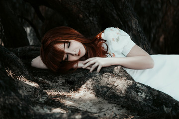 The little red-haired girl in a white dress walks in the mystical and mysterious forest.Princess with a crown standing near the tree,creative computer colors,fashionable toning © kharchenkoirina
