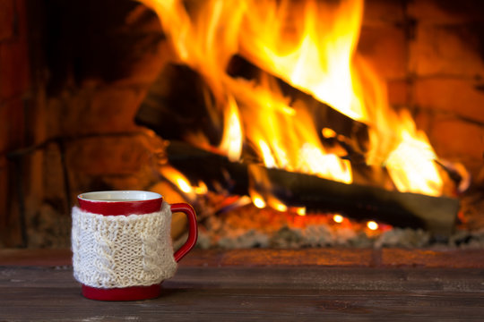 
Cup of hot drink and antique books in front of warm fireplace. Red mug in white knitted mitten standing near fire. Magical relazed cozi atmosphere.
