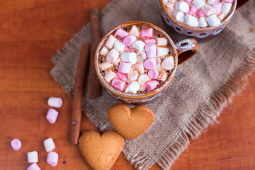 
Coffee and hot chocolate with marshmallows in a cup of pink and white with cinnamon and cookies in the shape of a heart on wood background
