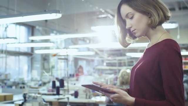 Young Woman Is Standing In A Clothing Factory And Using A Tablet While Employees Work In The Background. Shot On RED Cinema Camera.