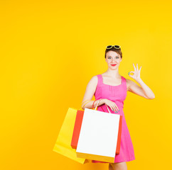 Smiling beautiful woman with shopping bags showing OK  on yellow background