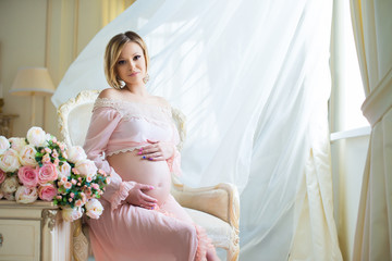 Cute pregnant woman sitting on chair near billowing curtains and hugs belly with love