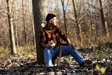 Portrait of senior lumberjack in forest sitting on a tree stump. He is resting and having lunch break.