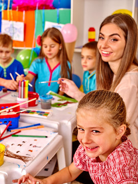 Large group of kids with female teacher painting on paper at table in kindergarten . 