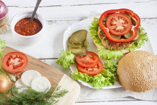 Homemade hamburger on white plate, sliced tomatoes, onion, pepper, lettuce and dill on wooden board, tomatoes  souse in ceramic plate. White wooden table.