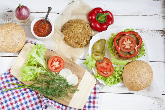 Homemade hamburger on white plate, sliced tomatoes, onion, pepper, lettuce and dill on wooden board, tomatoes  souse in ceramic plate. White wooden table.