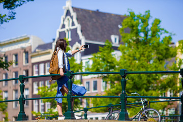 Obraz premium Young woman taking self portrait in european city outdoors. Young adult holding smartphone camera to take a picture of herself during her summer vacation in Amsterdam