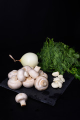 Vegetarian cooking ingredients: mushrooms, herbs, onion and garlic on stone board, black background.