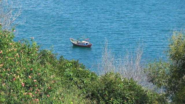 small wooden boat rocking on the waves. Bay is surrounded by greenery. Blue Sea has a slight wave.
