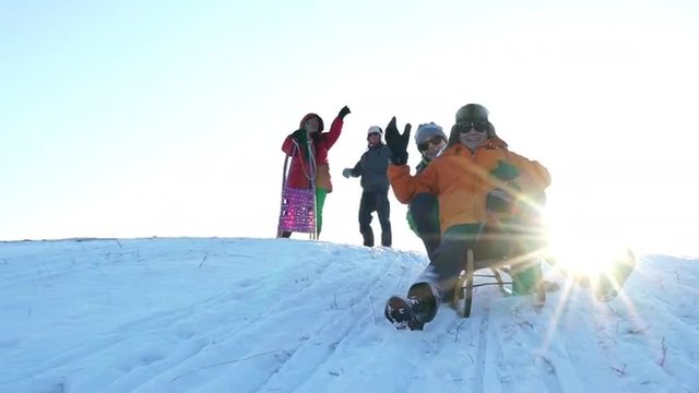 four active seniors having real fun on cold sunny winter day outdoors in snow, laughing best ager couple tobogganing while other couple waving on hill top, slow motion
