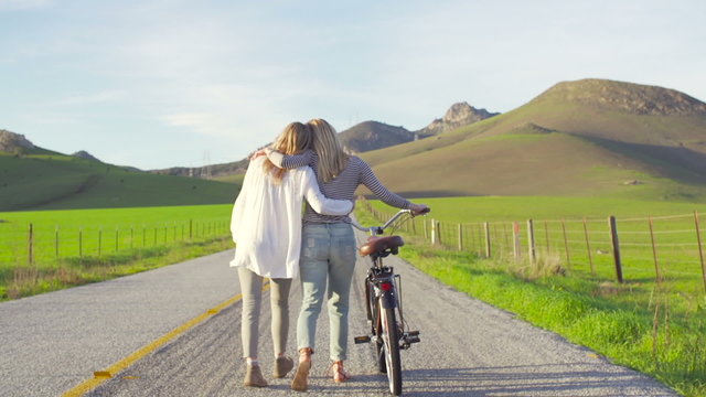 Two Girl Best Friends Walking Down Country Road With Bike At Sunset 