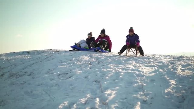 4k footage, three young girls tobogganing down snowy hill with sled and bobsled on sunny winter day
