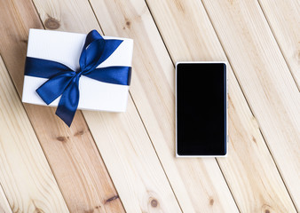 White Smart Phone With Blank Black Screen And White Gift Box With Blue Ribbon On Pine Table. 