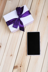 White Smart Phone With Blank Screen And White Gift Box With Violet Ribbon On Pine Table. 
