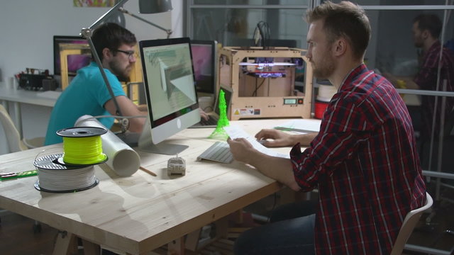 Two young men creating 3d models with computer software and printing them on 3D printer at small design studio 