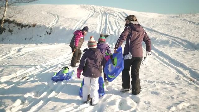 4k footage, following three childern with mother walking up snowy hill with bobsleds on sunny winter day
