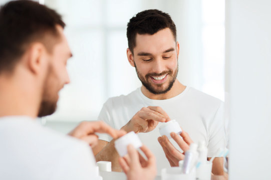 Happy Young Man Applying Cream To Face At Bathroom