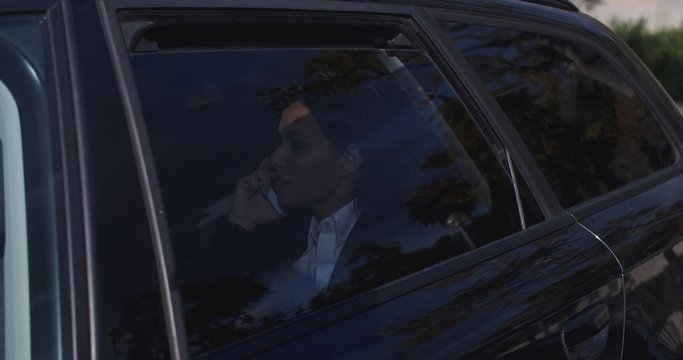 Serious young female executive with button collar on phone and sitting in rear seat of limousine with window down