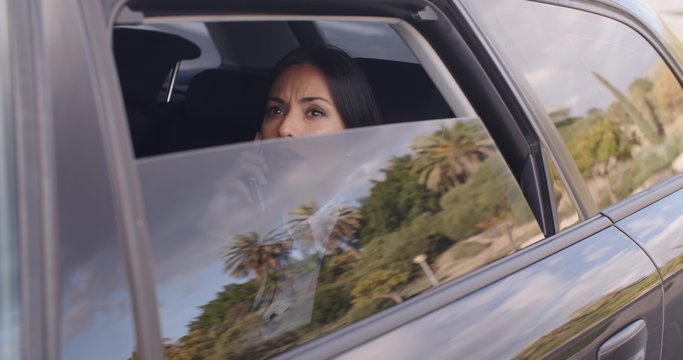Serious young woman in business attire on cell phone while sitting in rear seat of limousine looking outside from open window