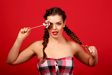 Studio close colorful portrait of a young sexy funny crazy fashion girl posing on a red background wall in style year dress with a pink lollipop.