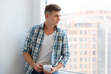 Handsome man sitting on windowsill and drinking coffee