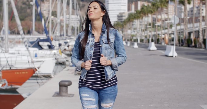 Trendy jaunty young woman enjoying a stroll along a waterfront promenade smiling as she looks out over the moored boats