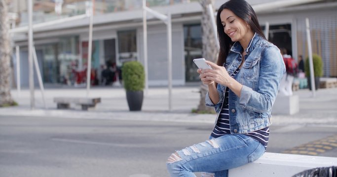 Trendy attractive young woman in a denim outfit perching on a wall on an urban promenade talking on her mobile and smiling at the camera
