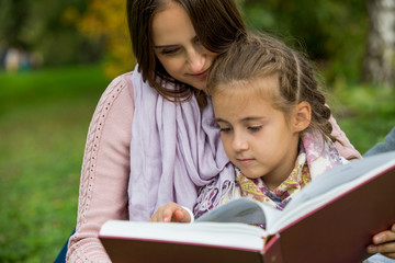 Young beautiful family sitting on the green grass in the park with big book. Reading fairy tale and smiling. Happy healthy family.