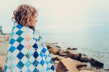 Fototapeta premium cute curly happy child girl relaxing on stone beach, wrapped in cozy quilt blanket. Summer vacation activities, traveling in Europe on holidays.
