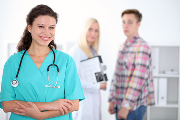 Beautiful young smiling female doctor standing at hospital with doctor and patient on the background. Medical concept.