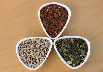Eatable seeds (sunflower, pumpkin, flax) in three bowls arranged in a three-leaf shape, top view