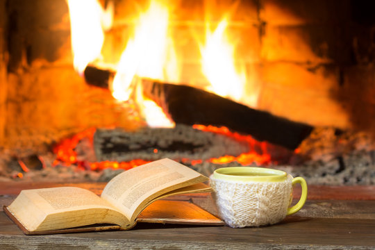 Cup of hot drink and antique books in front of warm fireplace. Yellow mug in white knitted mitten standing near fire. Magical relazed cozi atmosphere.