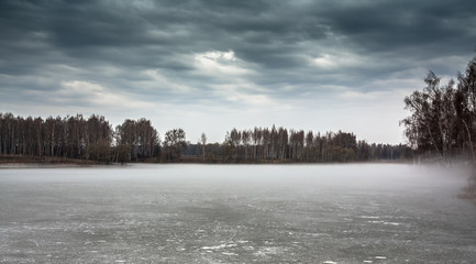 Gloomy landscape on frozen misty lake in season between winter and spring