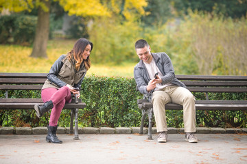 Two young people sitting on benches in a park and exchanging phone numbers