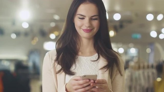 Happy young brunette girl is standing in a department store and texting a message on a smartphone. Shot on RED Cinema Camera.