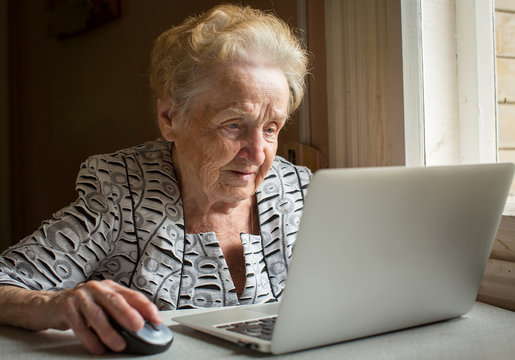 An elderly woman sitting at the table and types on laptop.