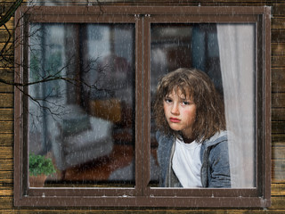 Rain, window of a house. Outside, the teen girl in the apartment. Girl sad, crying. The house is dark. Portrait of the girl in tears outside the window. Water drops on the window