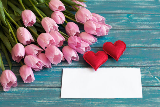 bouquet of pink tulips lie on natural wooden textur table, next to a white envelope and two red handmade hearts