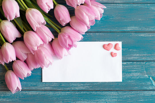 bouquet of pink tulips lie on texture painted in blue color table, next to a white envelope and three pink handmade sugar hearts