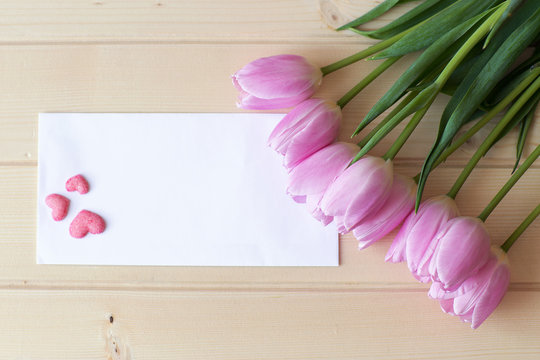bouquet of pink tulips lie on natural wooden texture table, next to a white envelope and and three pink handmade sugar hearts