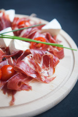 slices of prosciutto with mixed cherry tomatoes and cheese brie on wooden cutting board over the dark grey background. Flat lay concept 