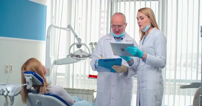 Dentist discussing diagnosis of his patient with young female assistant, they using the tablet then looking at camera and smiling