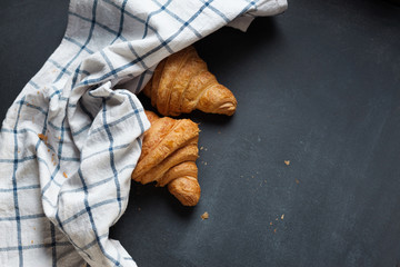 Two fresh croissant lying on a blackboard. Also, there is twisted linen towel. Croissants are delicious and appetizing. Towel is white with blue pattern.