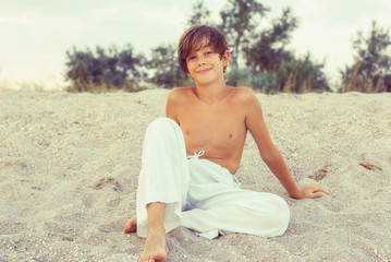 Smiling boy wearing a wide white pants, sitting in the sand on t