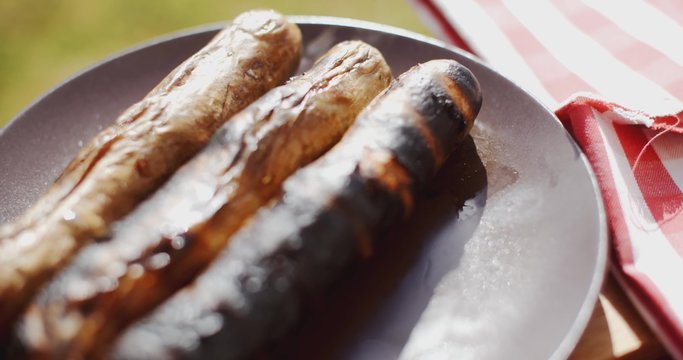 Row of three charred sausages on wooden board outside next to napkin with grass in the background
