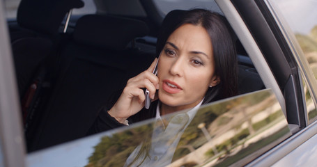 Serious young woman in business attire on cell phone while sitting in rear seat of limousine looking outside from open window