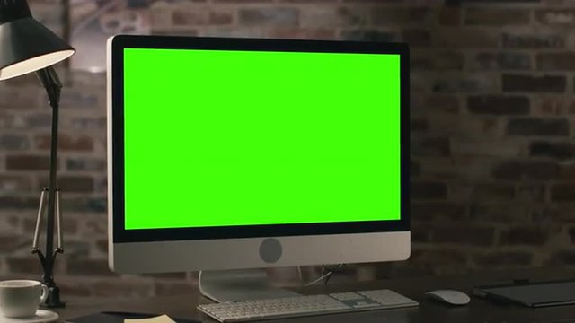 Footage of a computer monitor with green screen standing on a table next to a tablet, lamp, coffee cup, notebook and mouse in a loft. Shot on RED Cinema Camera.