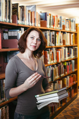 Portrait of middle age mature brunette Caucasian woman student with glasses in library holding book, looking directly in camera, teacher librarian profession, back to school concept